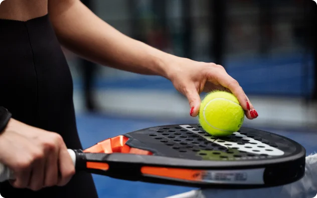 Gros plan sur une personne tenant une raquette de padel dans sa main gauche et plaçant une balle de padel jaune sur sa surface. L'arrière-plan est un court intérieur flou